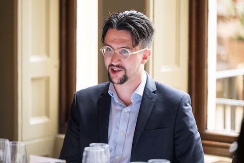 Photograph of Insurance Times deputy editor Yiannis Kotoulas. He wears clear-framed oval glasses and a light-blue shirt with a navy blazer over the top.